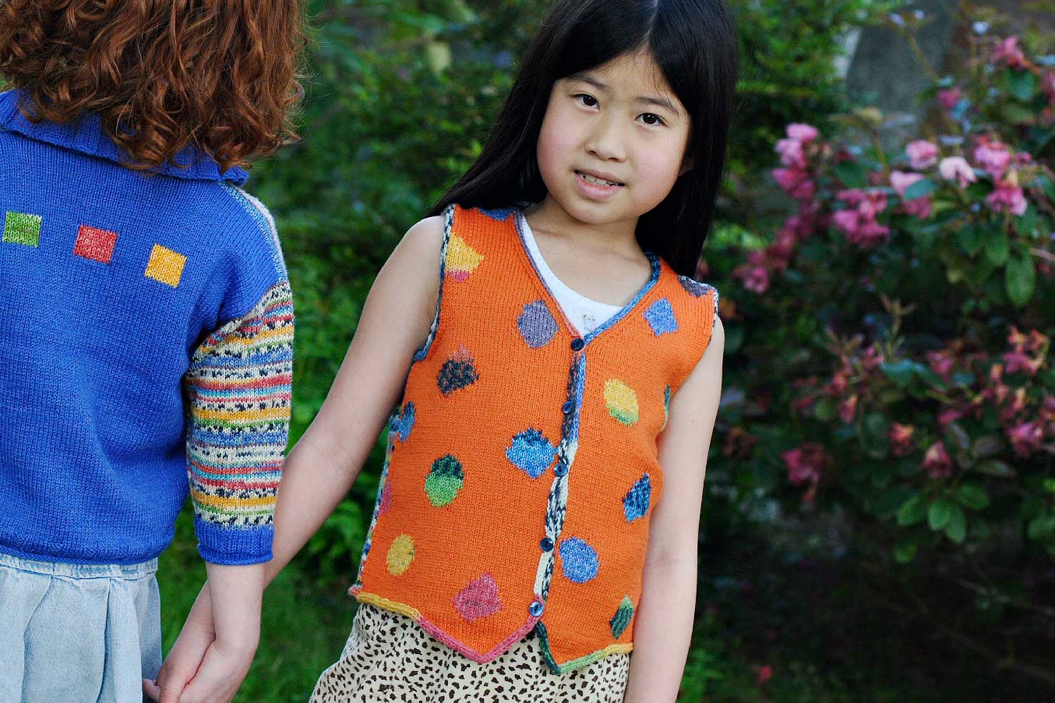 Two girls standing side by side holding hands and wearing bright sweaters.