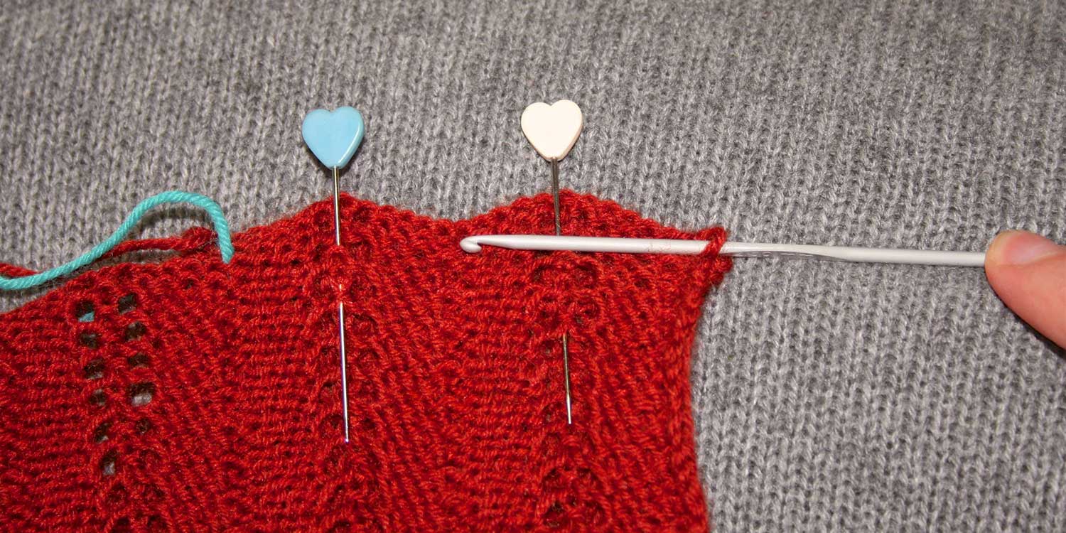 Wrong side of knitting showing with two pins; the bind-off edge is scalloped; a crochet hook has one stitch on it and is held horizontal, drawing a line across the knitting showing where to stitch a straight line.