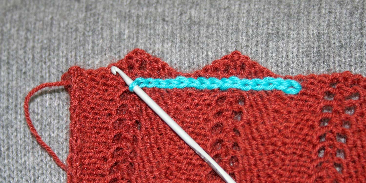 A turquoise slip stitch seam being worked with a crochet hook on a rust piece of knitting with a zig-zaggy top edge.