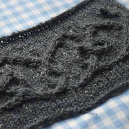 Swatch of cable motif on grey yarn, sitting on blue and white gingham fabric.