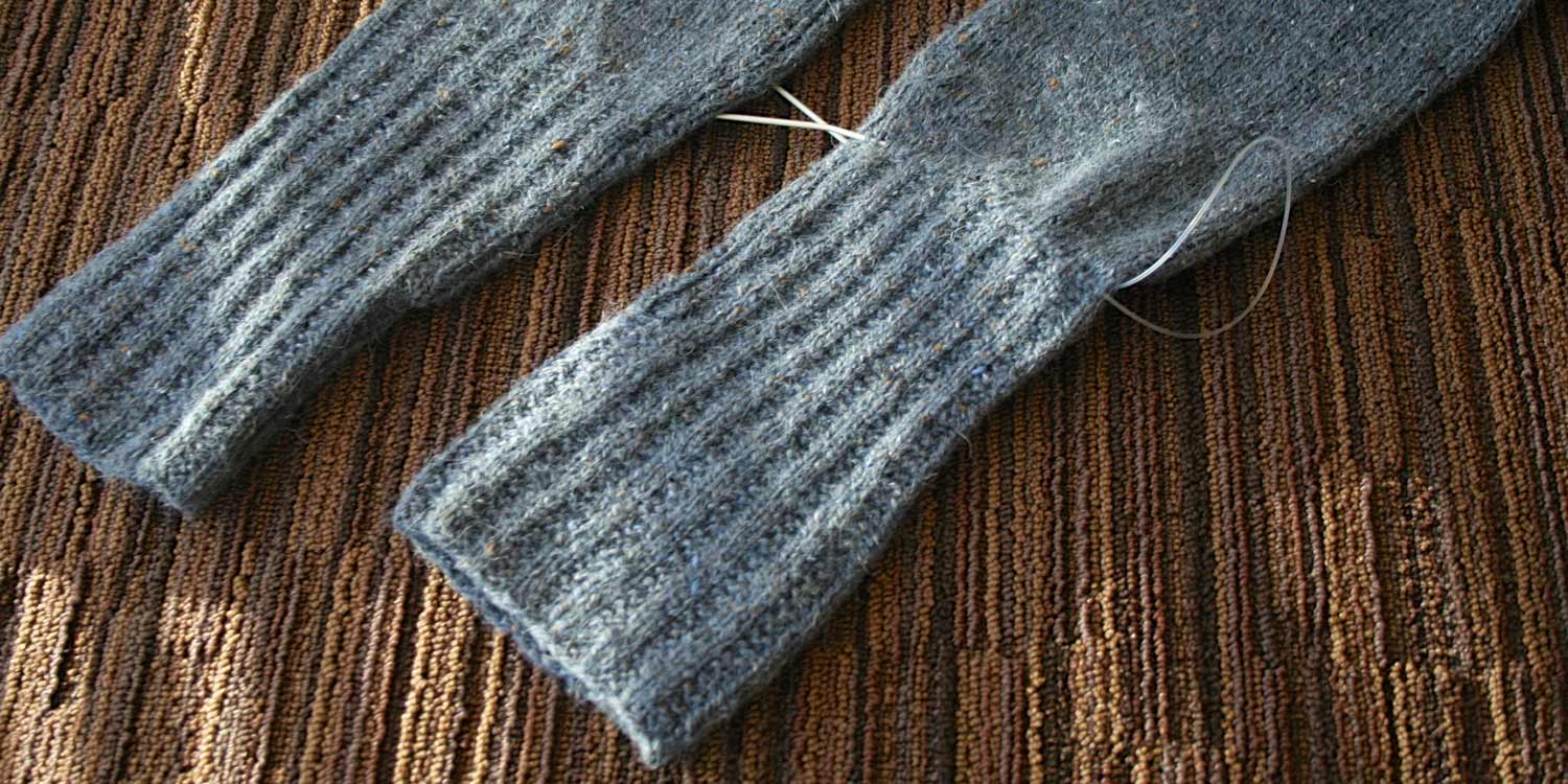 Two sleeve cuffs, one of which is oddly shaped and has a circular needle threaded through all the stitches just above the ribbing.