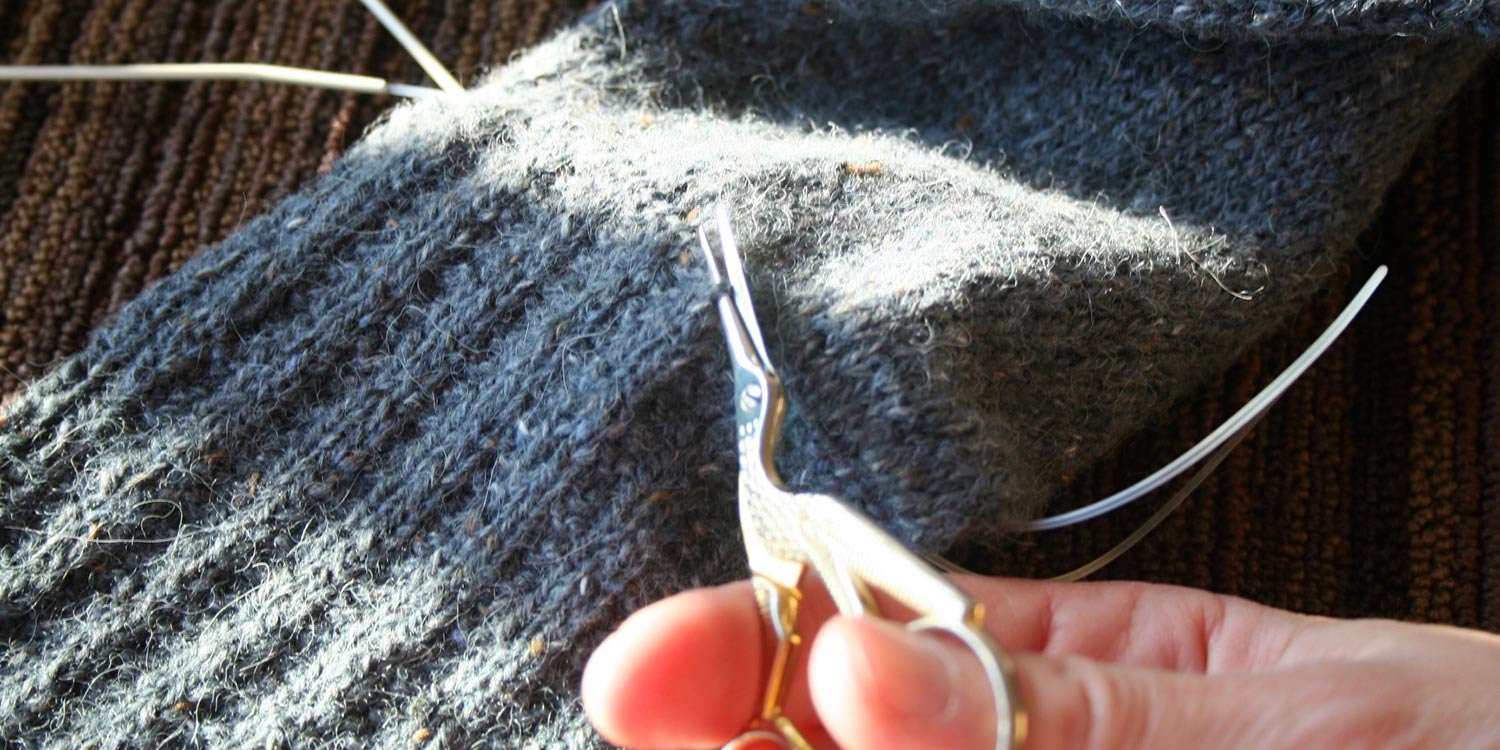 Hand holding a pair of stork-shaped scissors about to cut a stitch in some knitting.