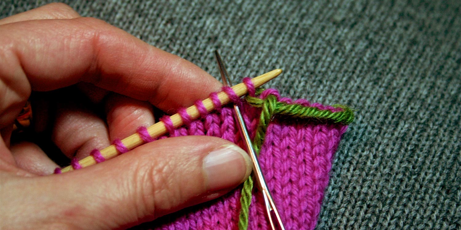 Threaded tapestry needle is inserted in second stitch on left needle; a partially bound off edge can be seen to the right.