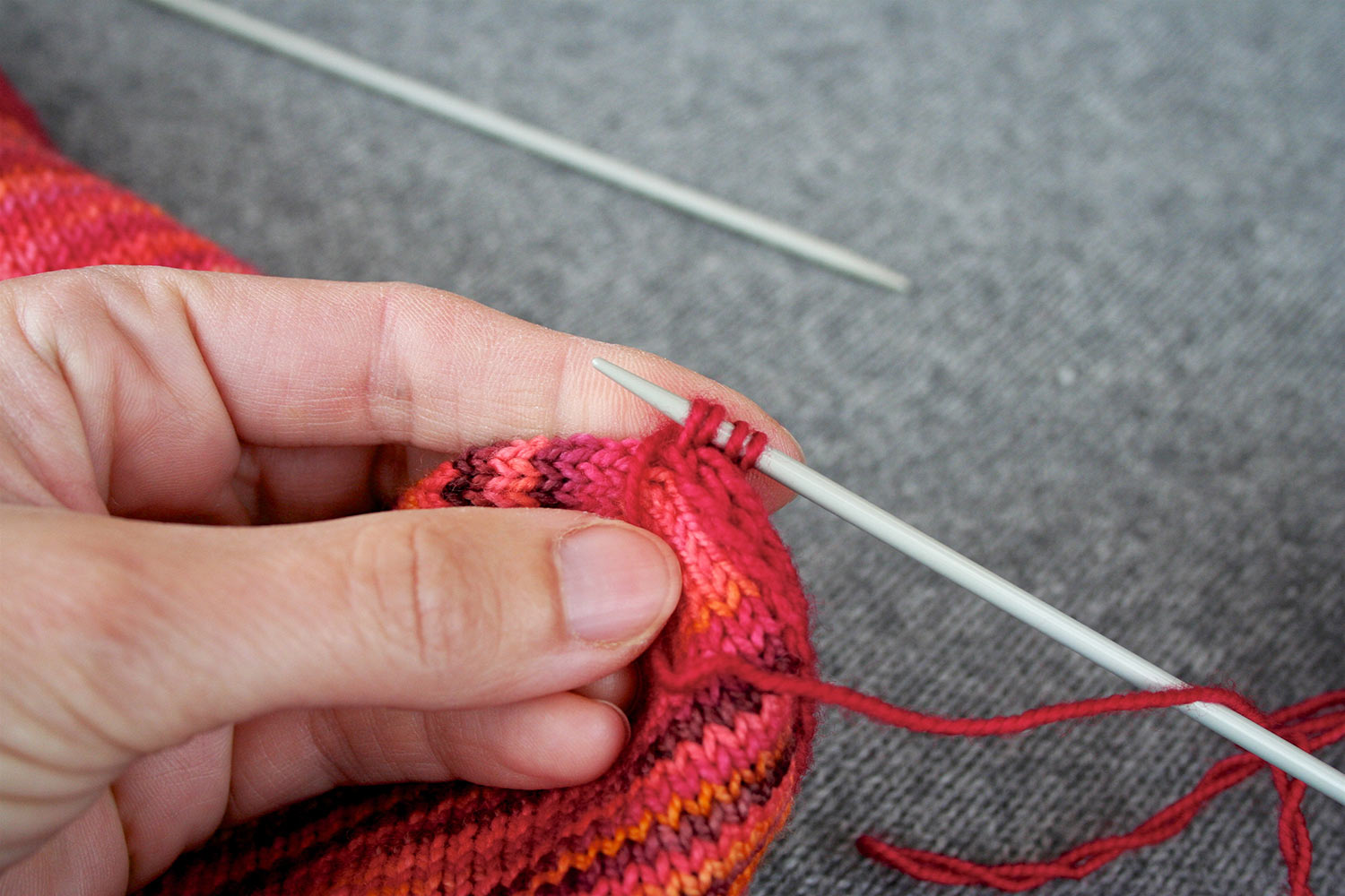 Four stitches on right needle with yarn brought to front and held in place by thumb of left hand; empty needle out of focus in background.