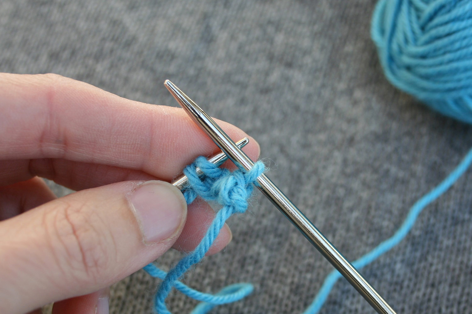 One stitch remains on right needle with a clump of stitches just below it created by casting on and then binding off a few stitches; left needle still has remaining cast on still on the needle.