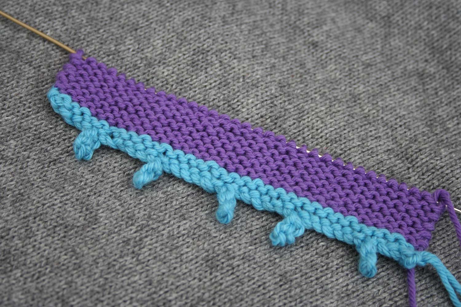 Knitting on the needle with a blue picot cast on with long picot bumps and six stitches between them.