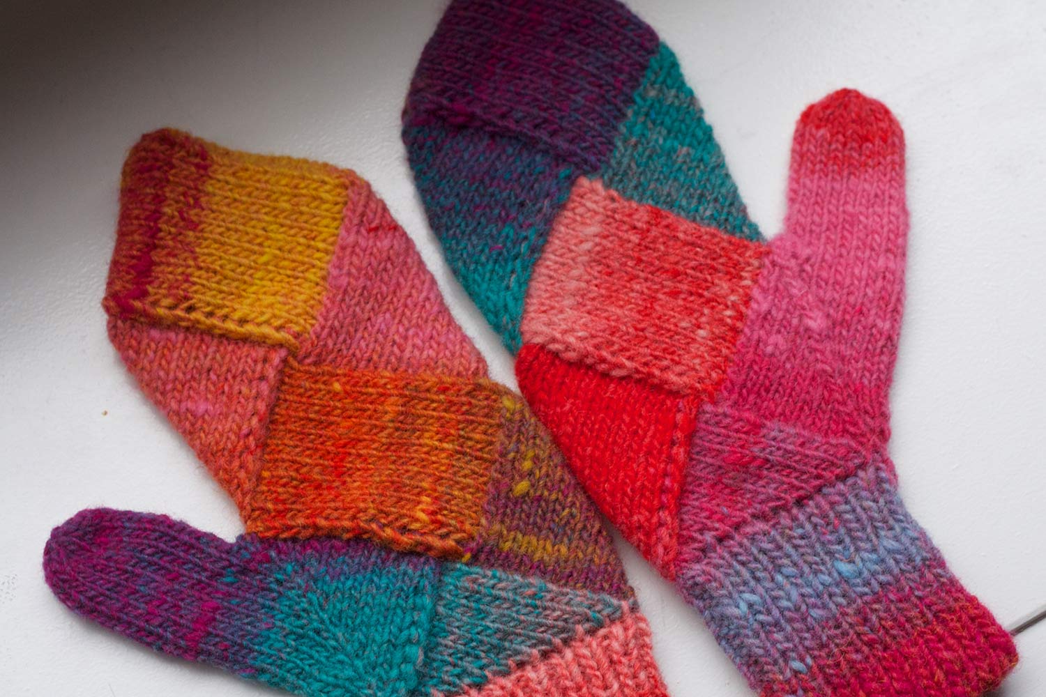 Colour mittens with thumbs pointing out on white surface; triangles and diamond-shaped blocks of colour worked in entrelac.
