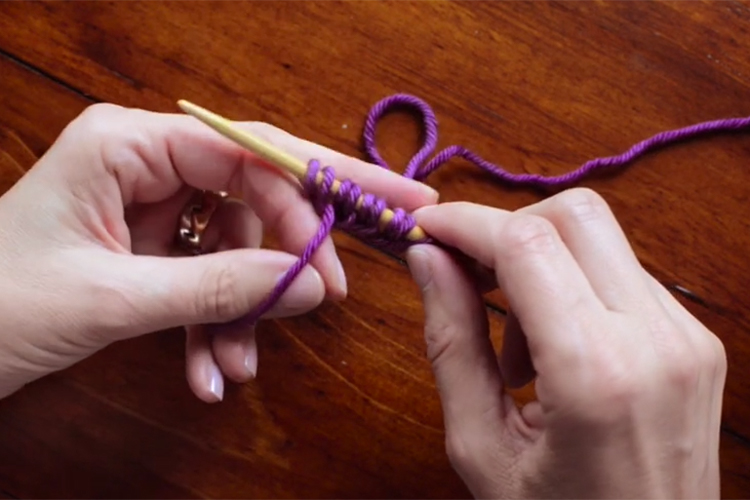 Knitting needle in right hand with a few stitches cast on and left hand is manipulating the yarn strands into a long tail cast on set up.