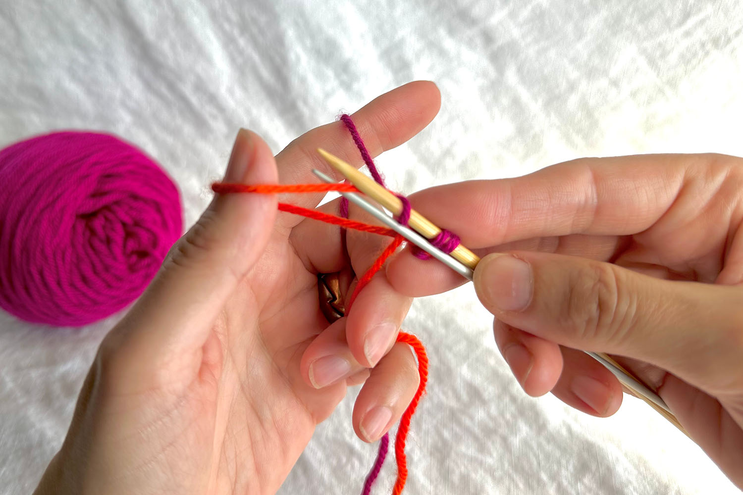 Near strand of thumb loop is brought between both needles and wrapped downwards around smaller needle.