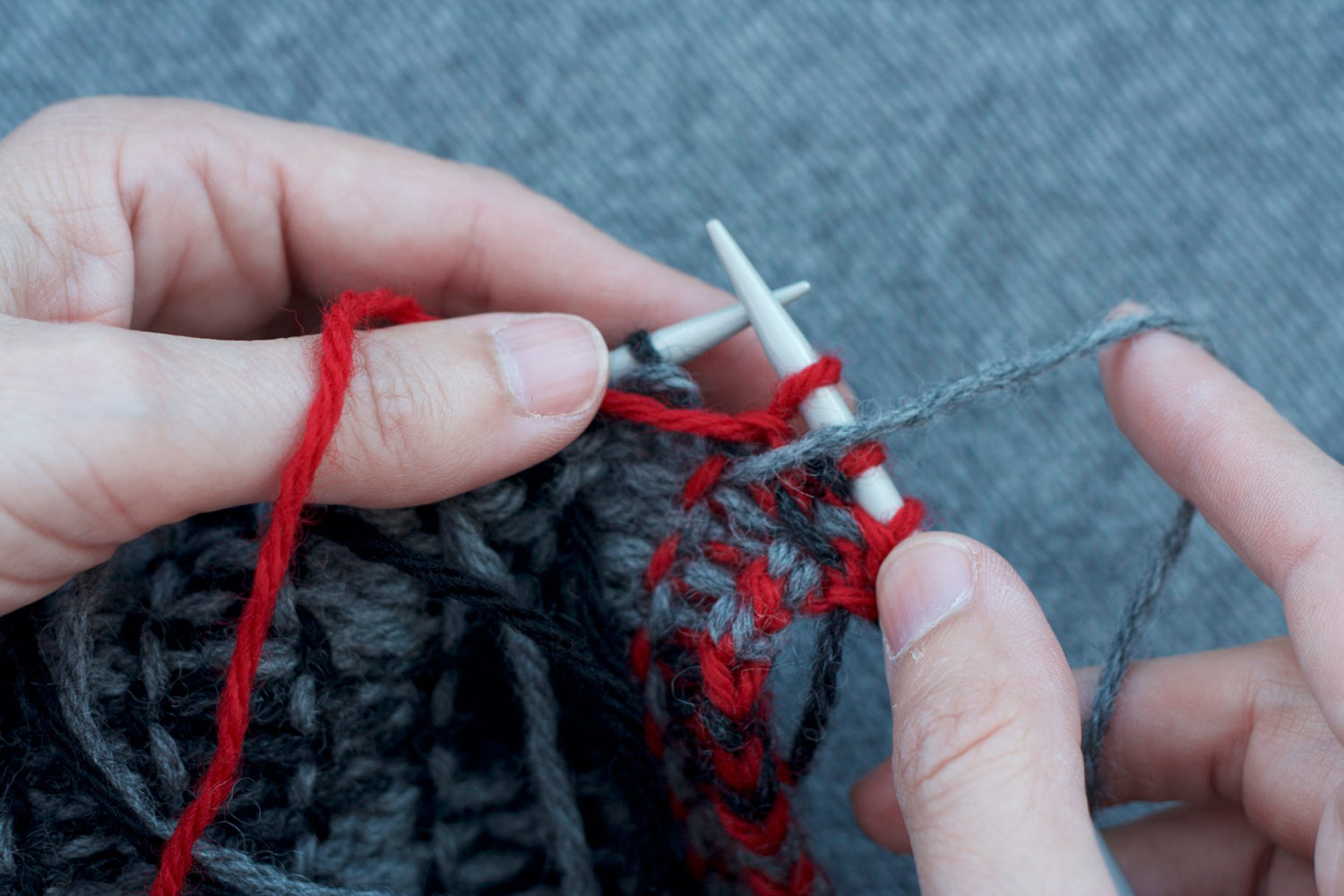 With the wrong side facing, hands show how to transition from red stitches to grey stitches in the intarsia technique; red yarn is held to the left and grey yarn is pulled up and to the right.