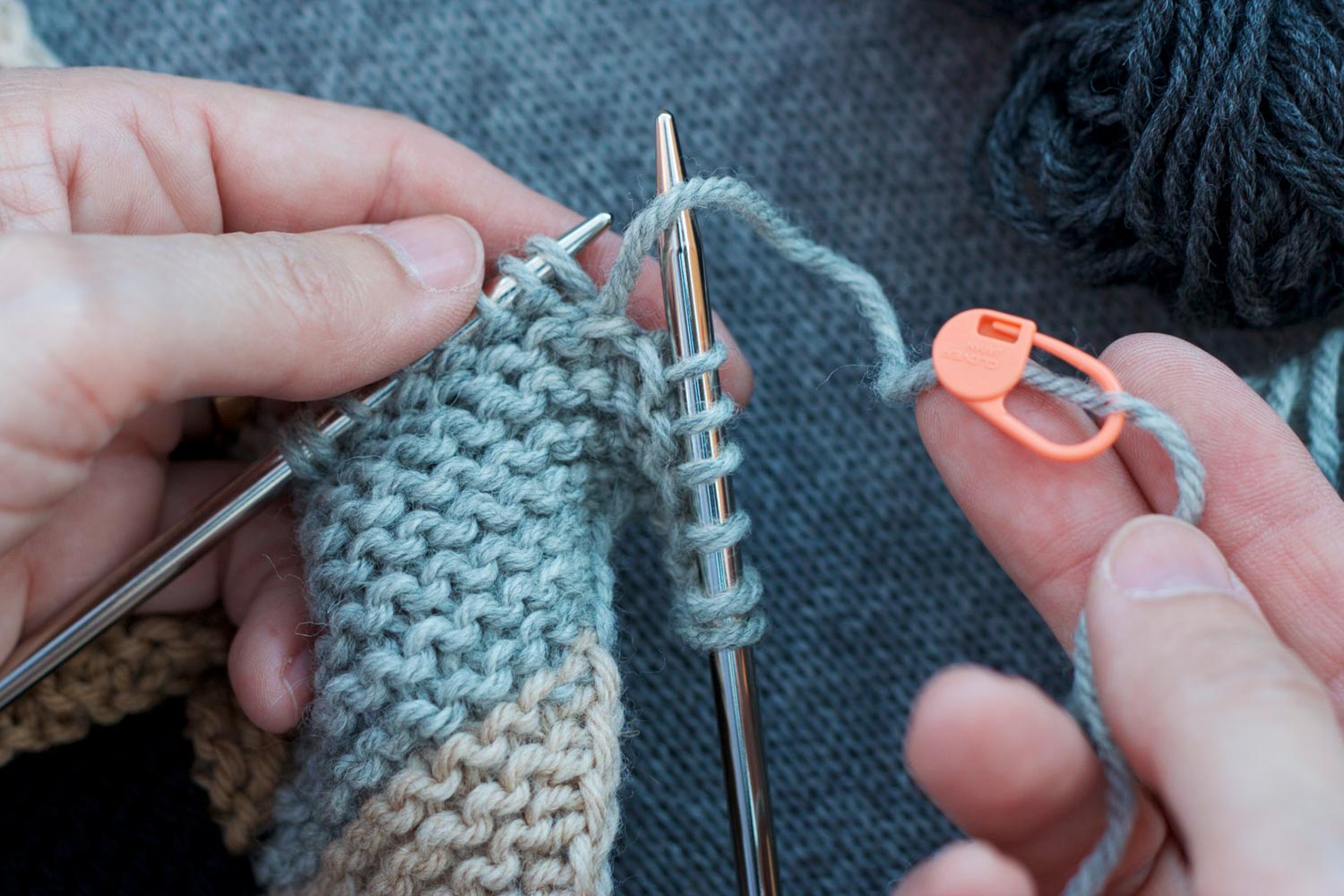 Six stitches unraveled on right needle and remaining stitches on left needle; right hand holds unraveled yarn with a stitch marker pinned several inches down its length.