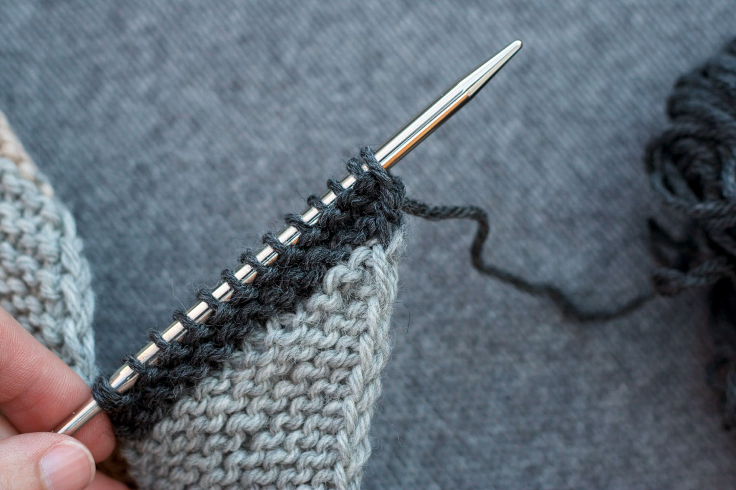 Several rows of light grey garter stitch with two charcoal grey ridges above, on the needles, showing that the two-colour join occurred perfectly at the end of a row.