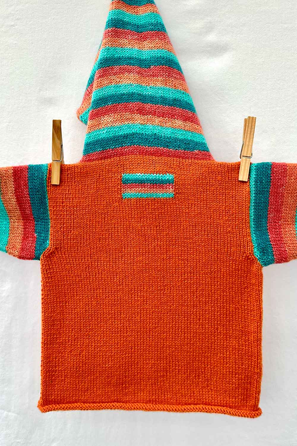 Back view of child's hoodie with striped hood, sleeves, and rectangular intarsia patch worked in self-striping yarn, hanging on a clothes line.