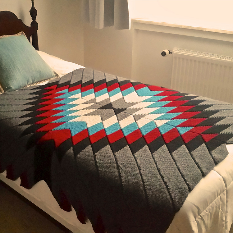 Modular knit garter stitch blanket on bed with geometric stylized sunburst in centre.