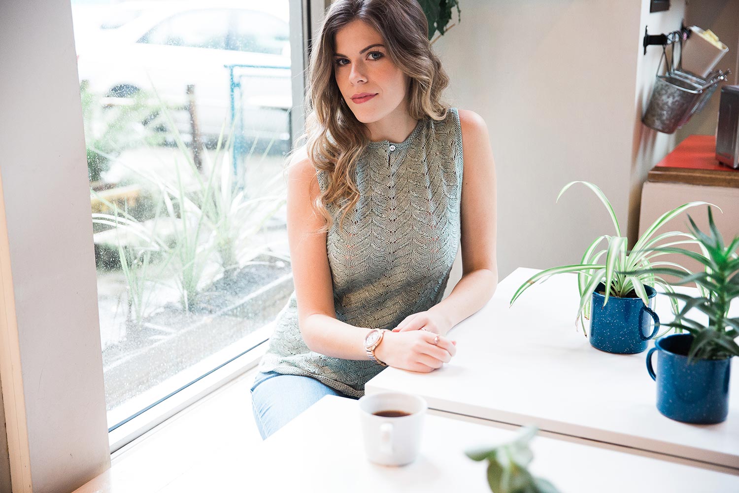 Seated woman facing camera with arms on table wearing sleeveless shell with allover lace pattern on front and an open button closure at neck.