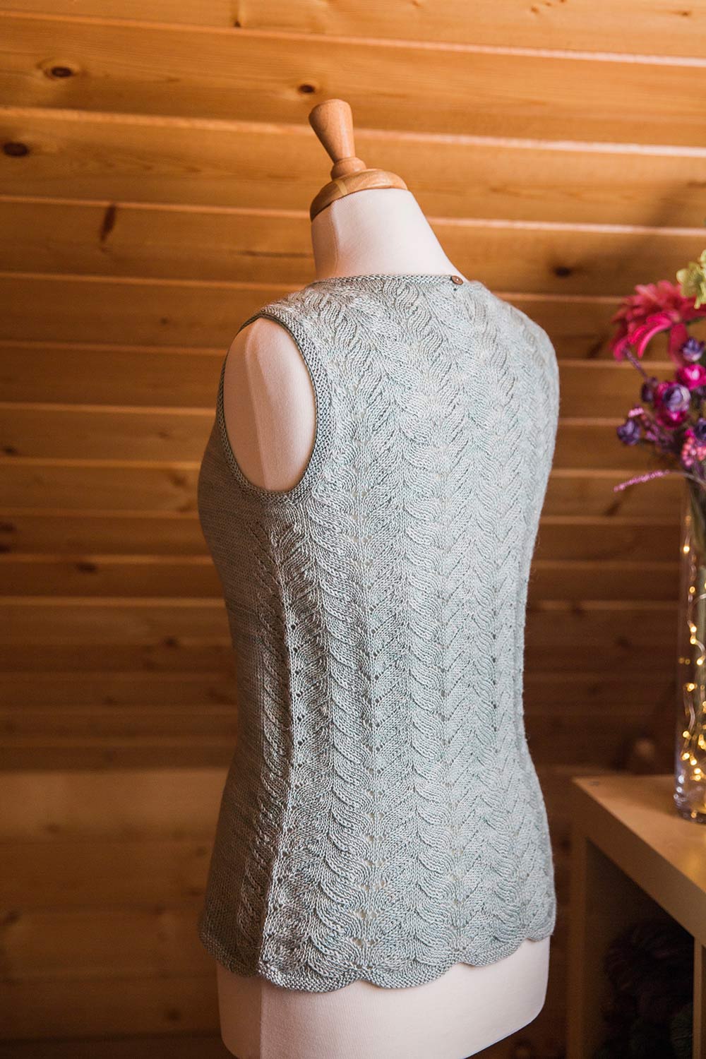 Back view of sleeveless shell on dress form with allover lace pattern on back, scalloped hem and a button closure at back neck.