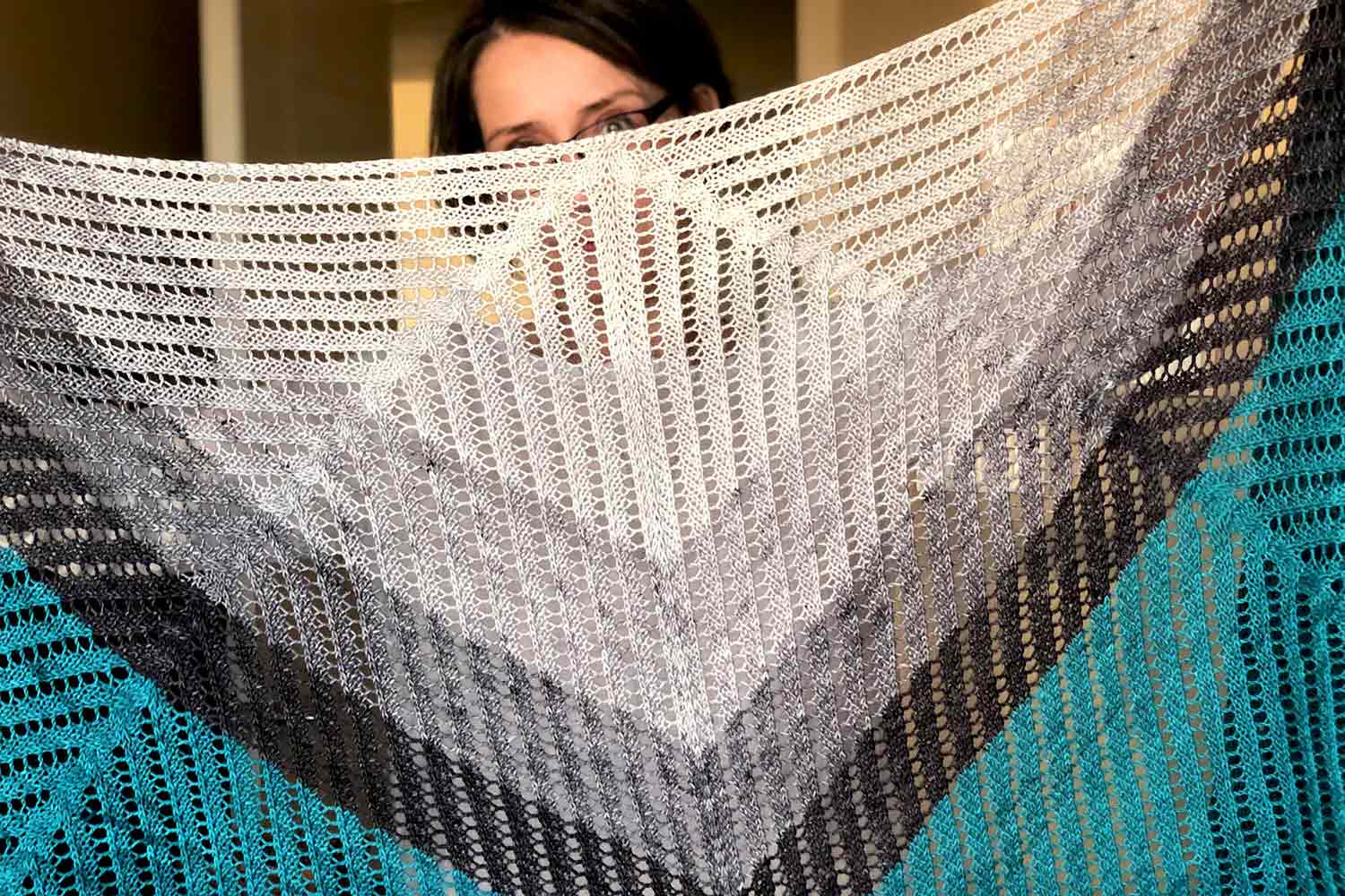 Holli peeking from behind the shawl she's holding up. Angles of eyelet lines are traveling against the bands of colour that form the central triangle of the shawl.