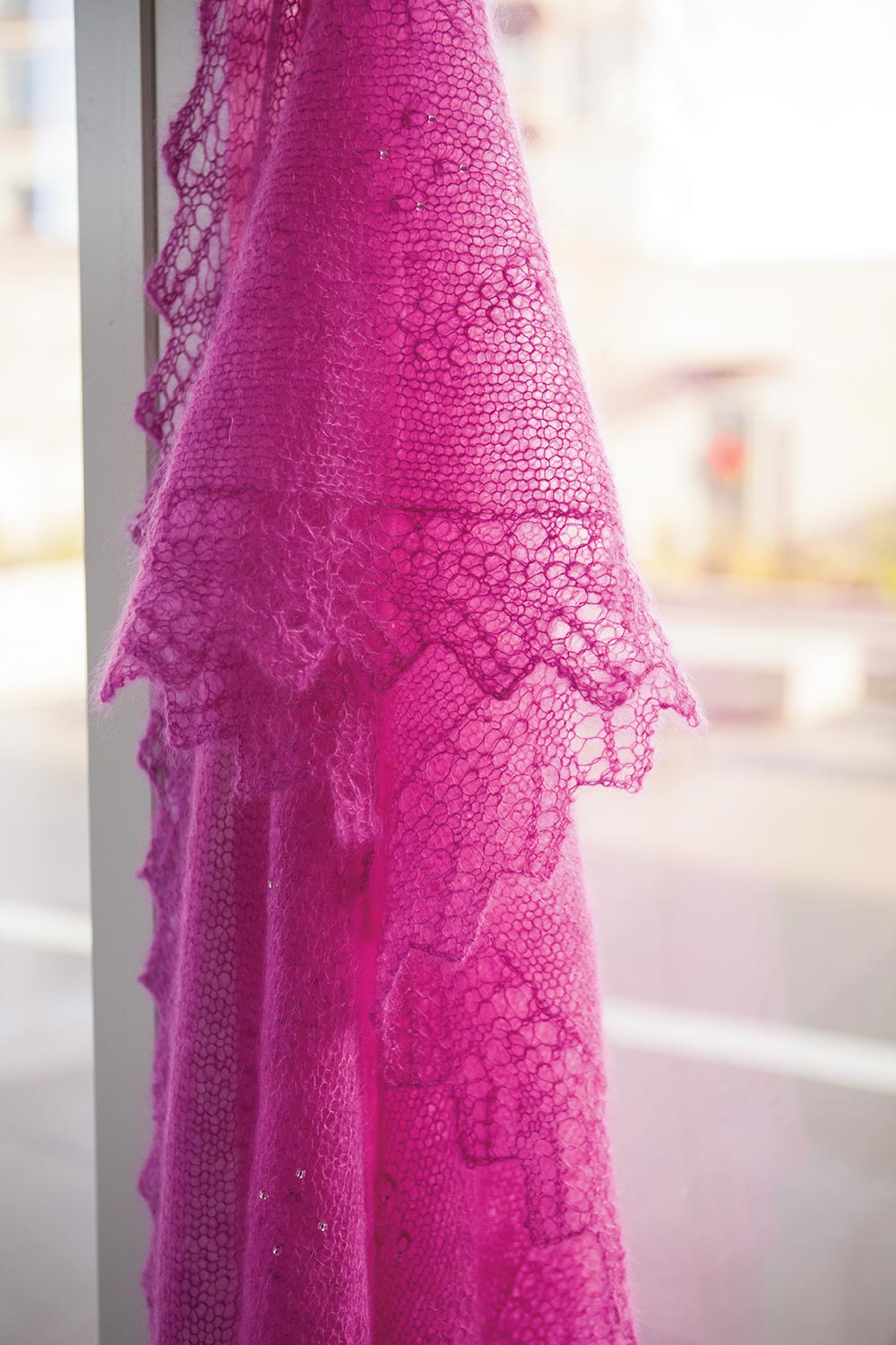 Pink mohair shawl is held up and allowed to drape, highlighting the delicate lace edging and showing some of the tuck stitch and bead patterning.