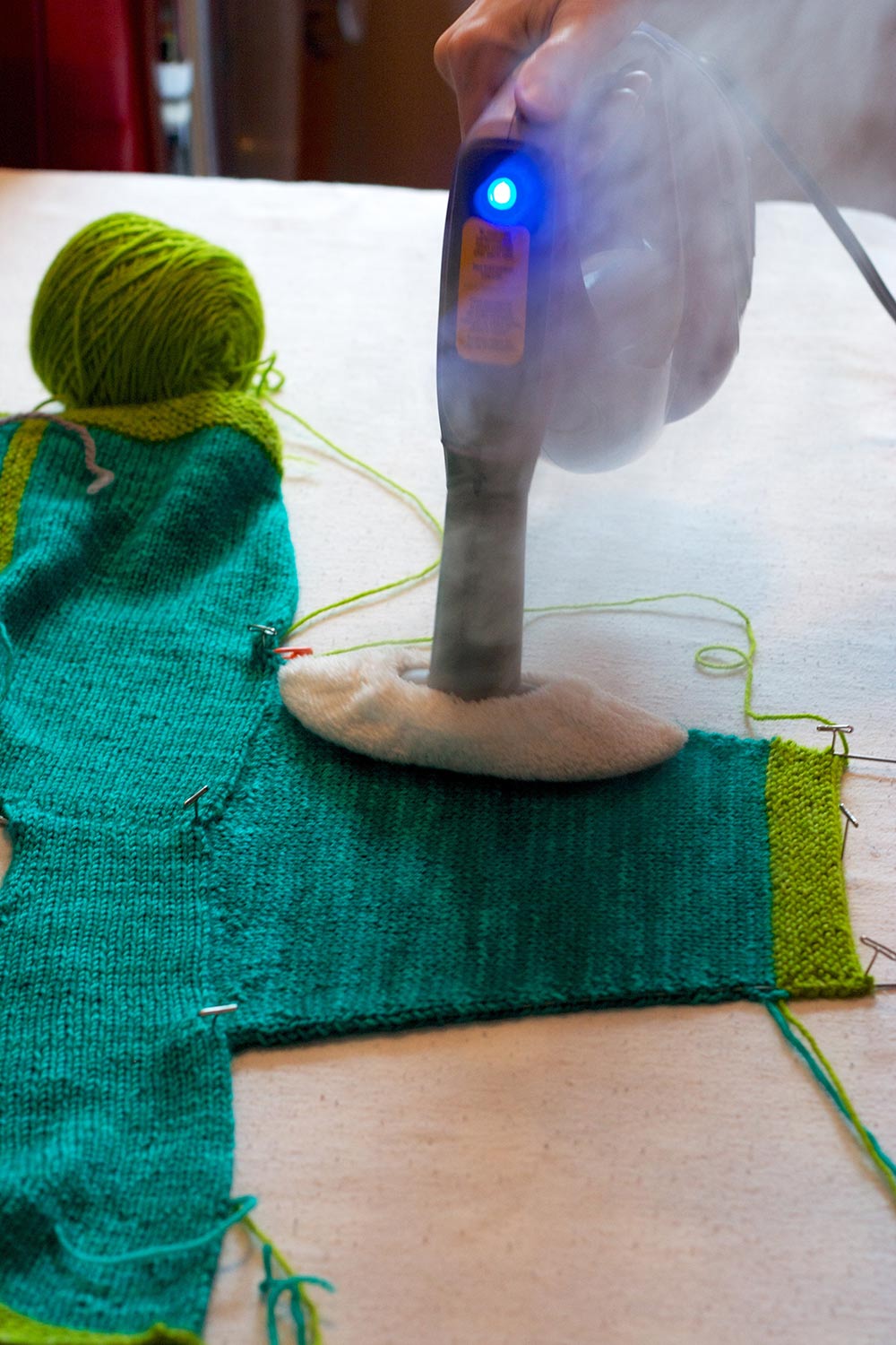 Partially assembled baby sweater laid out on blocking board and steam rising from hand held steamer.