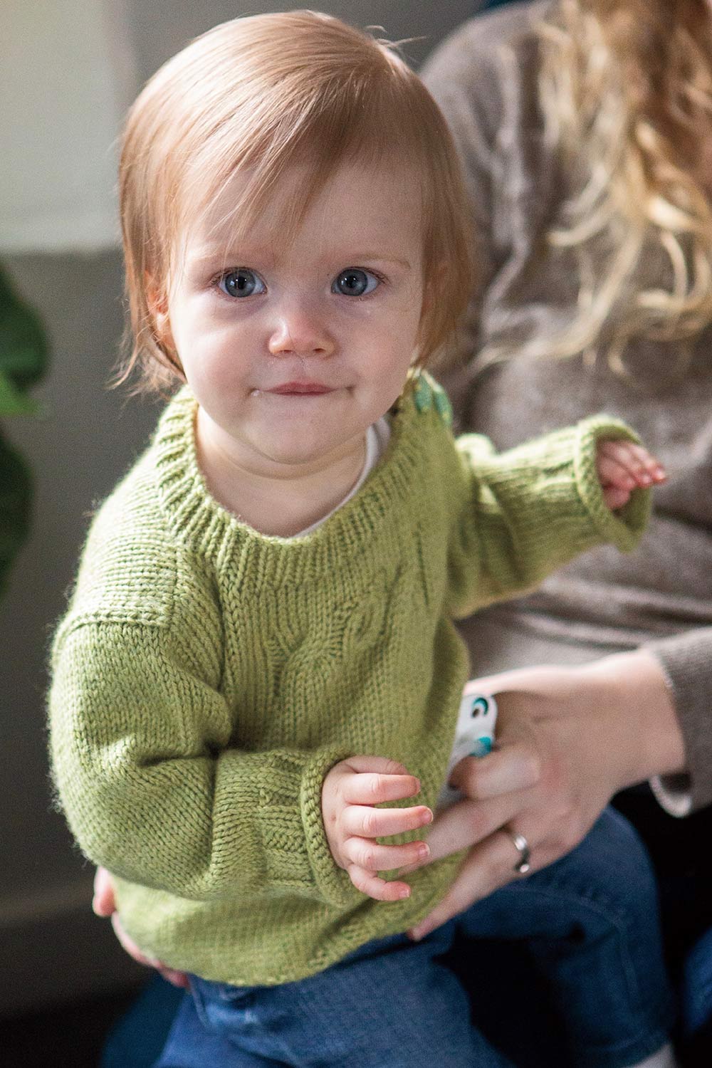 Baby in a stocking stitch pullover with ribbing at the neck and cuffs, a rolled cuff edge and cables on the chest.