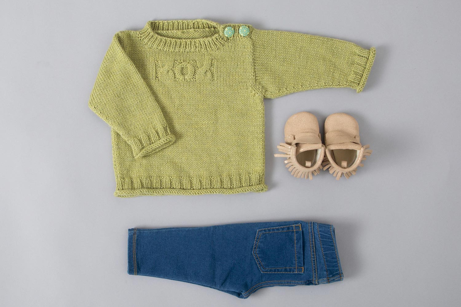 Flat lay of a light green baby sweater with buttons on the shoulder an three cables on the chest spelling out XOX. Baby jeans and booties complete the composition.