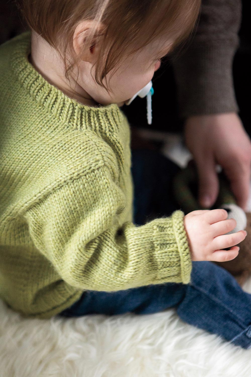 Shoulder and back view from above baby showing the ribbed neck and cuff of a stocking stitch pullover.