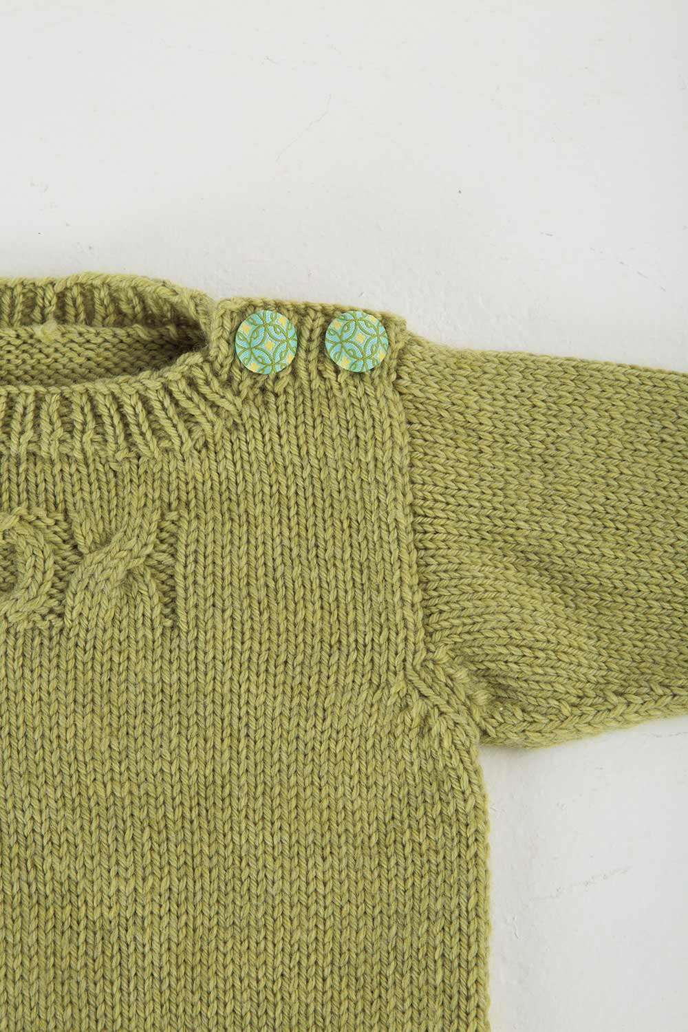 Detail shot of shoulder button placket with two buttons on baby sweater. An X cable and half of an O cable are visible on the chest.