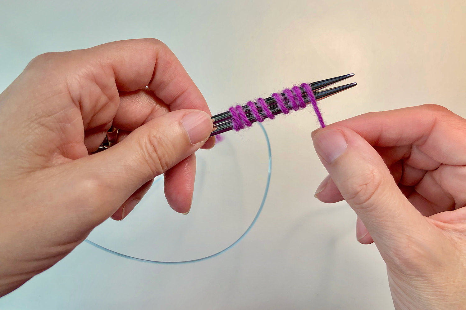 Hands holding two knitting needles together and wrapping pink yarn around them.