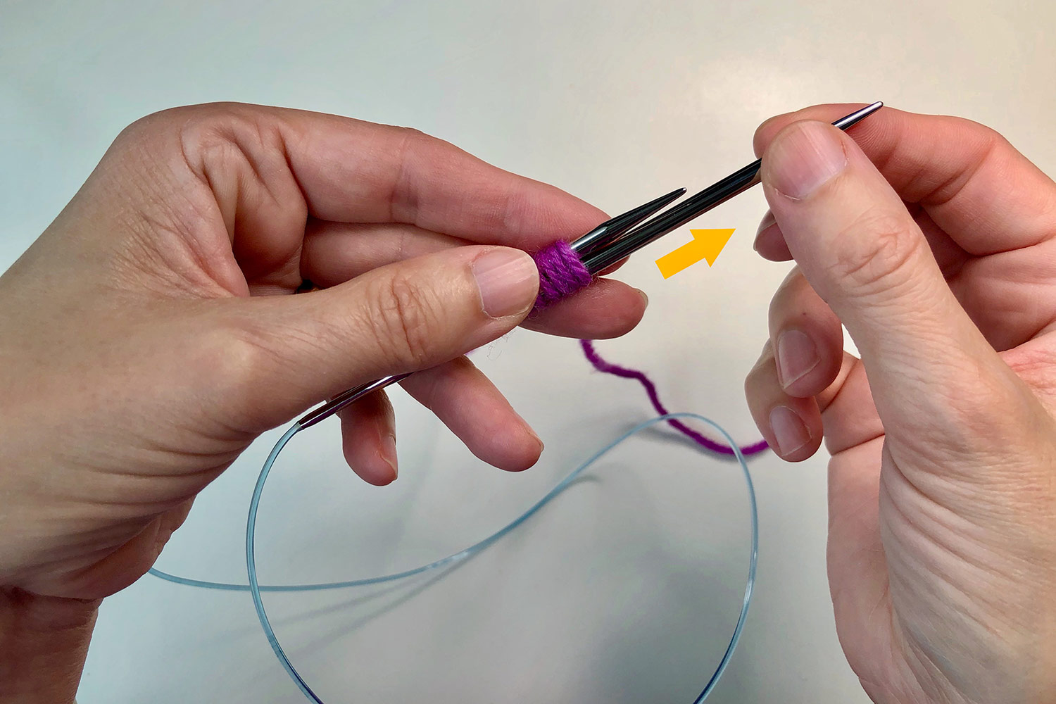 Yarn wrapped around two knitting needles with an arrow showing the direction to pull the lower needle.