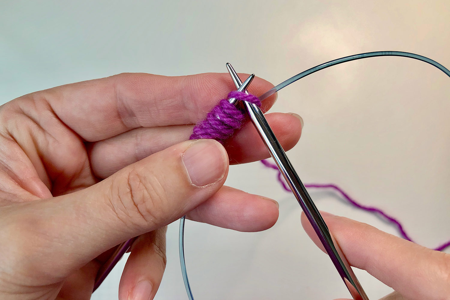 Pink yarn wrapped around a circular needle and its cable in the left hand with the other needle held in the right hand.