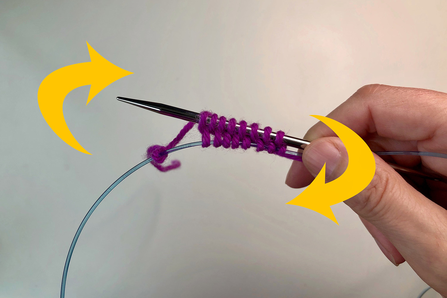 A small number of stitches on a needle with the cable part of the needle holding loops below the stitches; big yellow curved arrows showing the direction to turn the knitting.