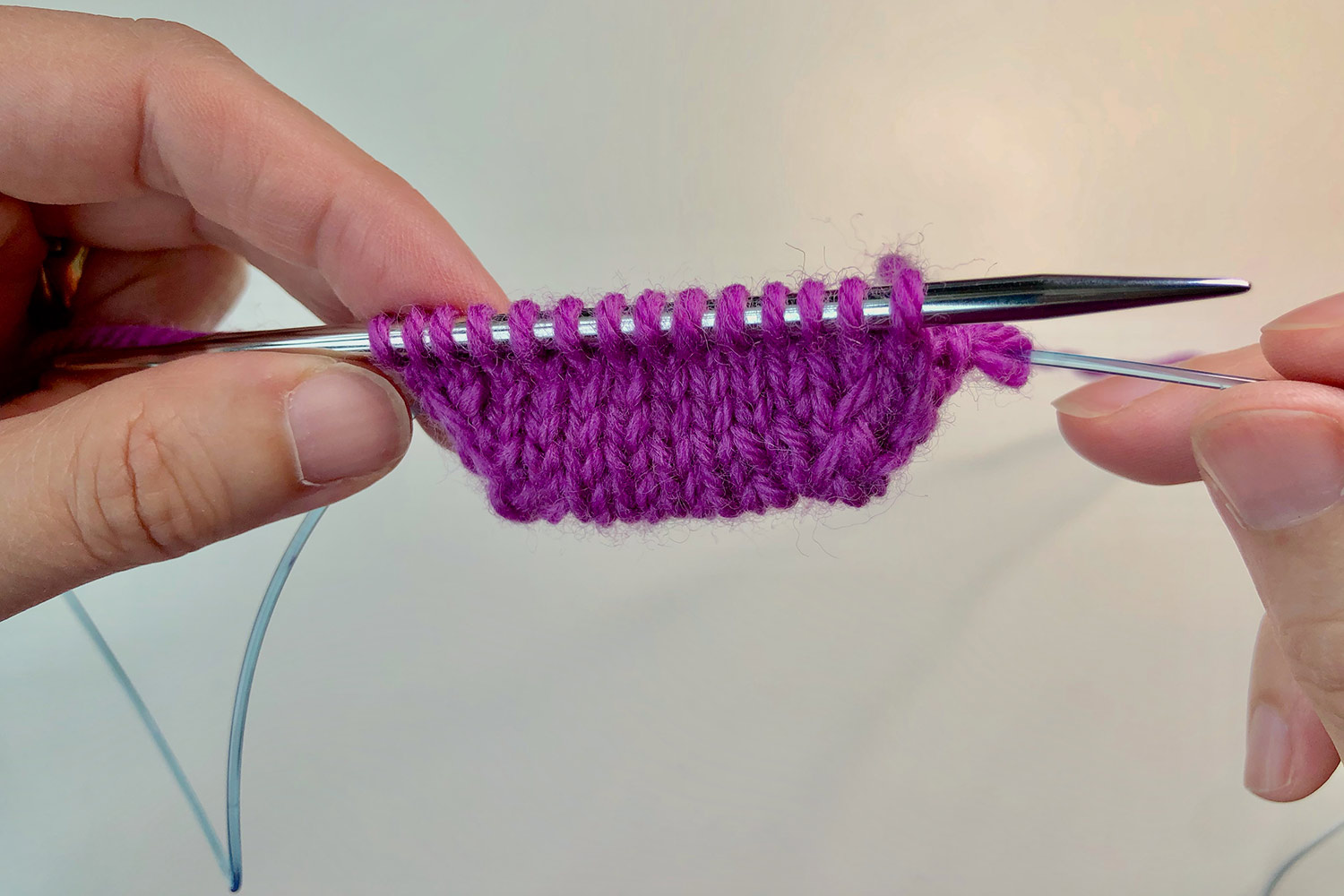 The beginning of a knitted toe on a needle and its cable behind held by two hands.