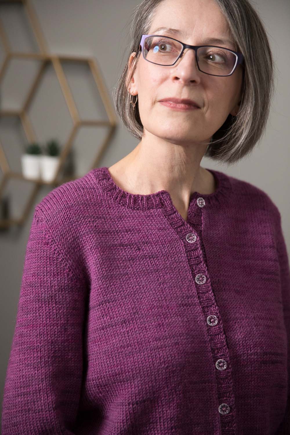 Woman wearing round-necked cardigan in purple hand-dyed yarn; all but the top transparent decorative button secure the opening.