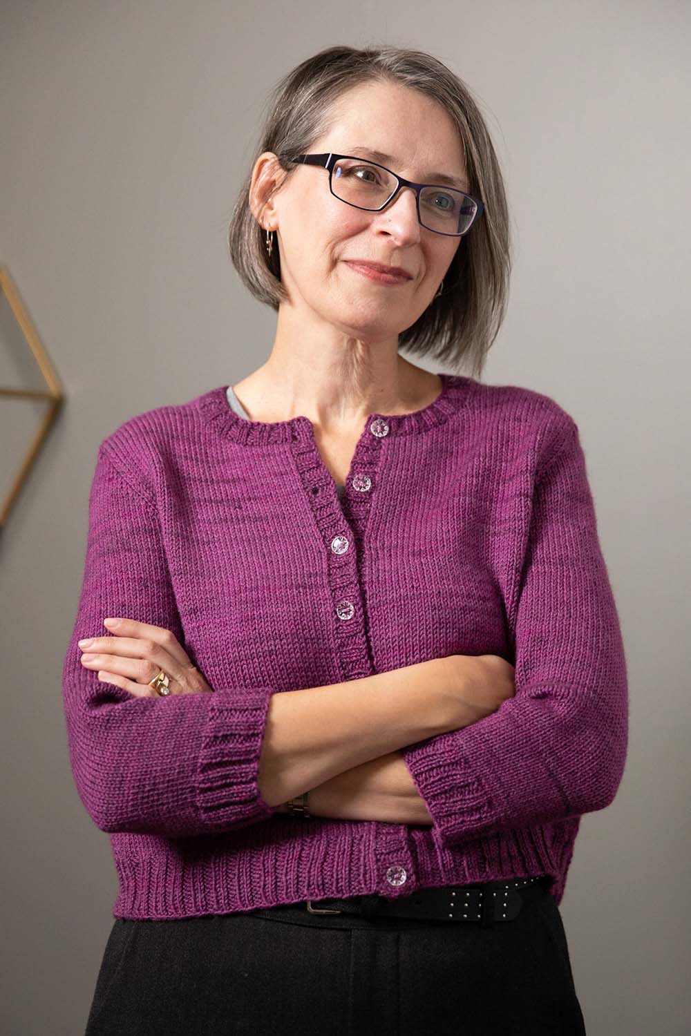 Smiling woman wearing round-necked cardigan in purple hand-dyed yarn; crossed arms show three-quarter length sleeves.