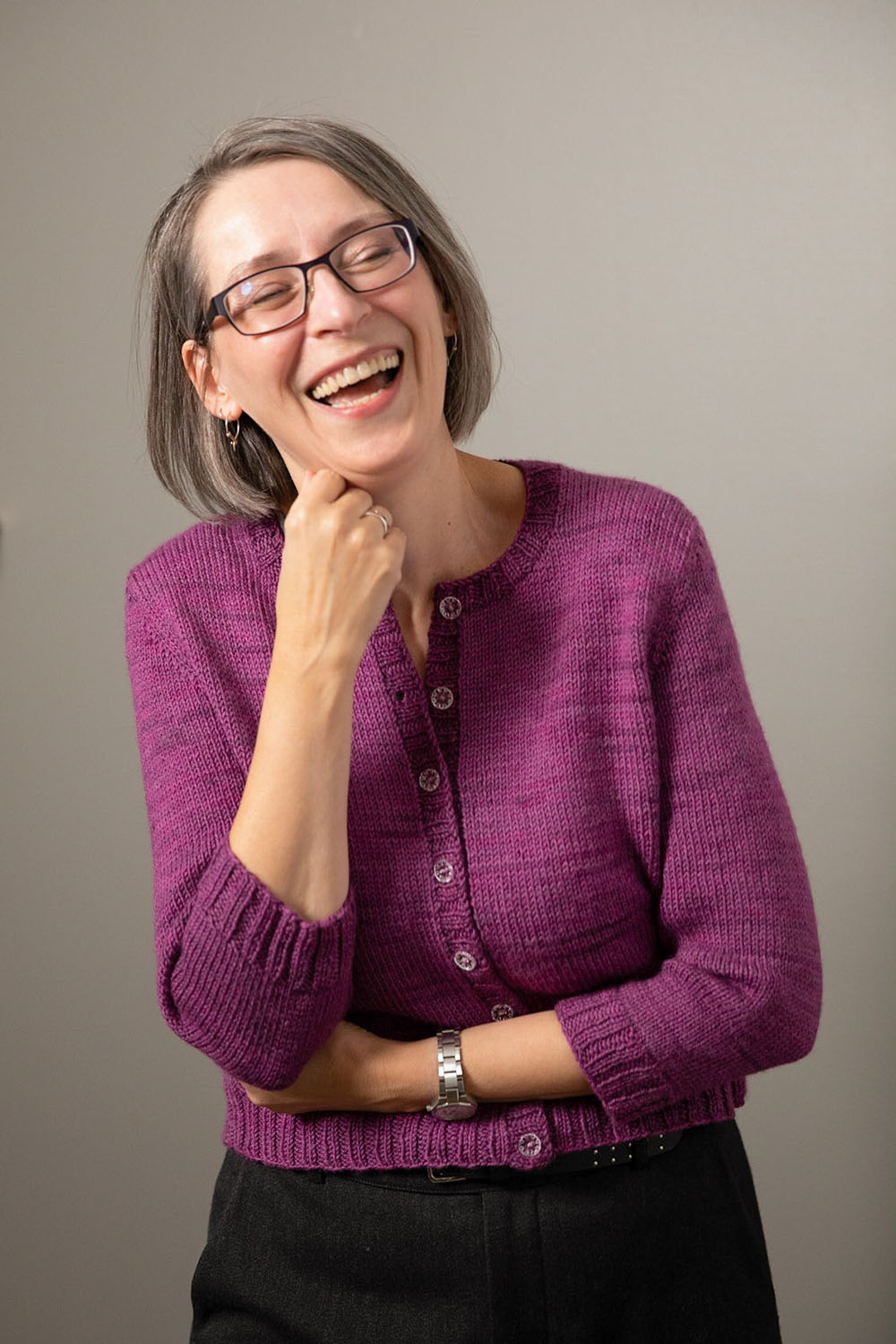 Laughing woman wearing round-necked cardigan in purple hand-dyed yarn; one arm crossed in front and other reaching up to chin shows three-quarter length sleeves.