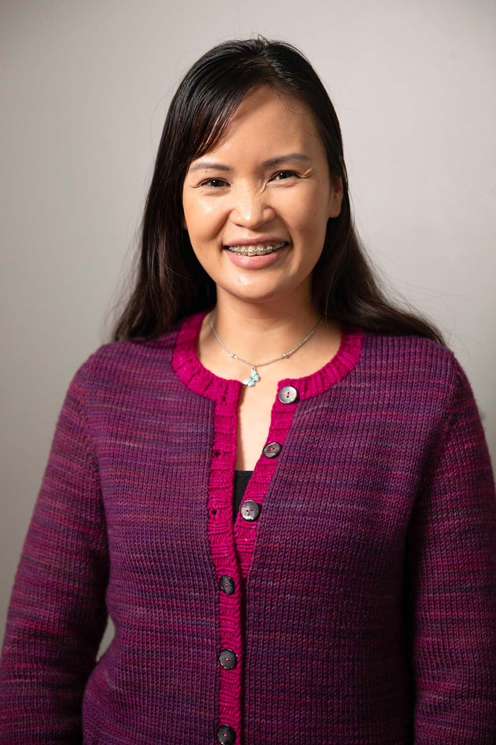 Smiling woman wearing round-necked cardigan in purple hand-dyed yarn with bright pink button bands and neck band.