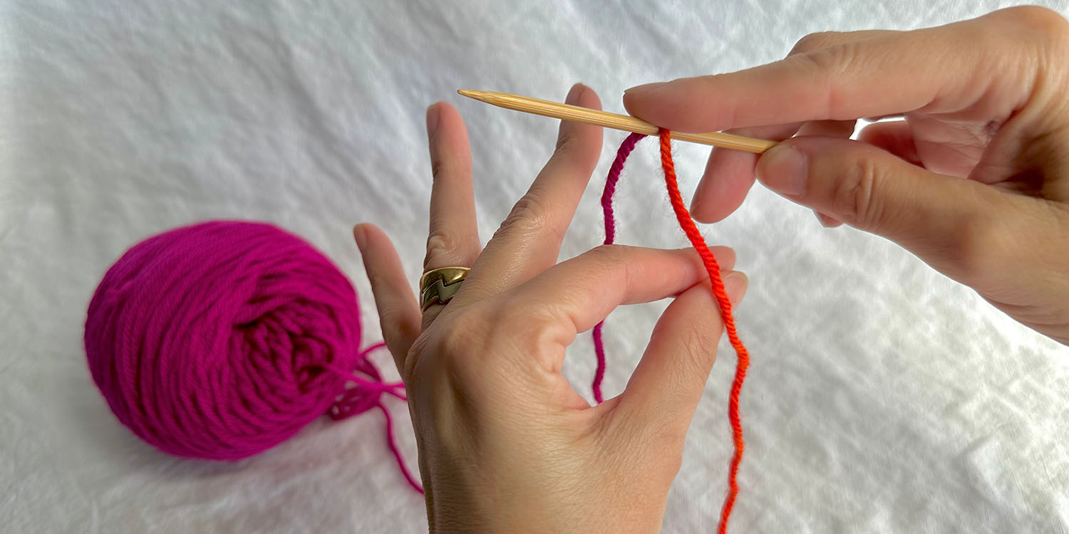 Thumb and index finger pinched together and inserted between two strands of yarn draped over needle.