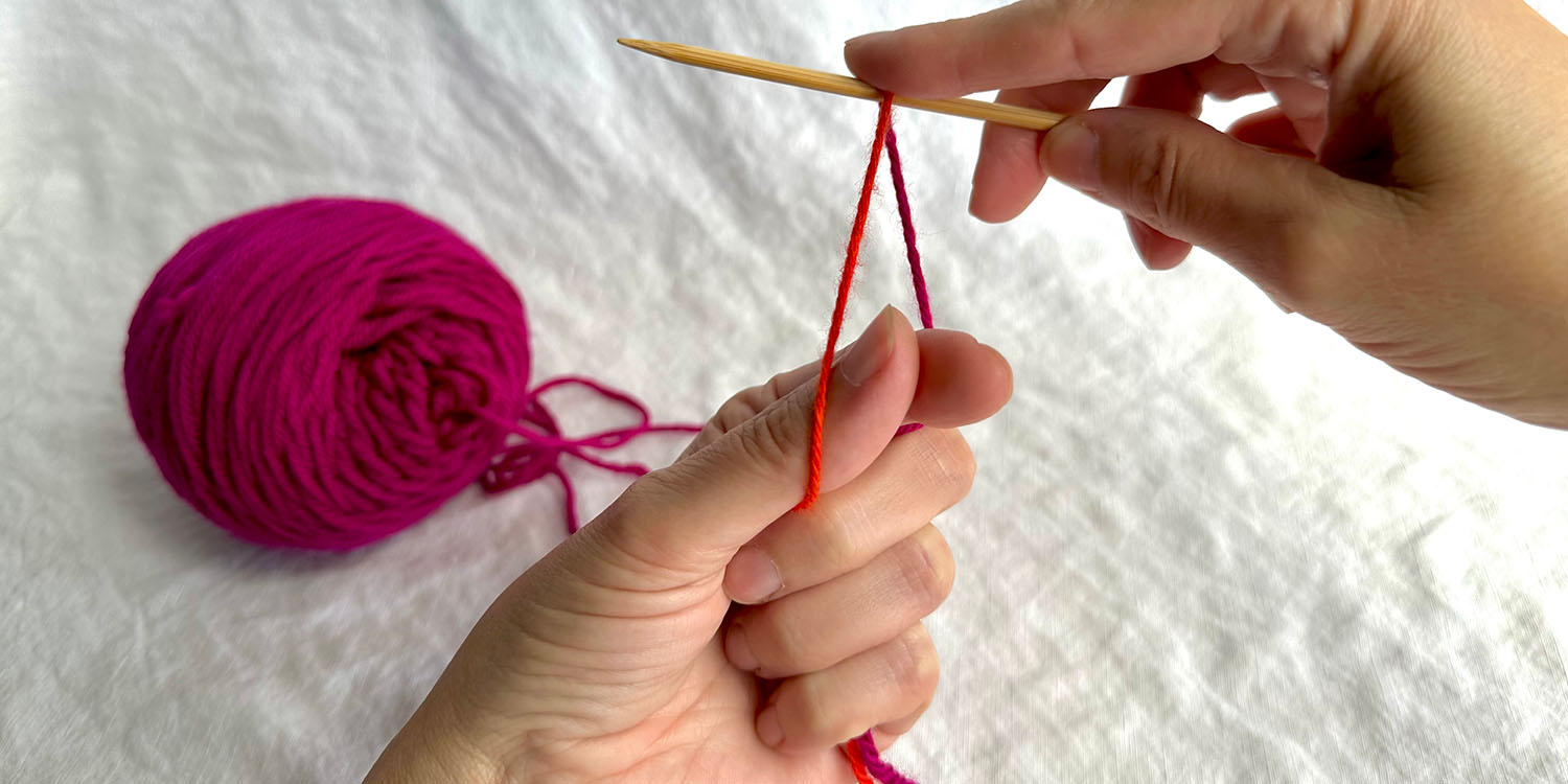 Two strands of yarn draped over needle and held in fist with thumb and index finger between the strands.