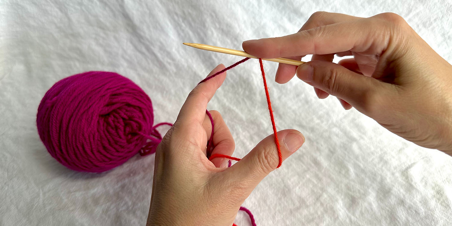 Thumb and index finger spread wide between two strands of yarn draped over needle.
