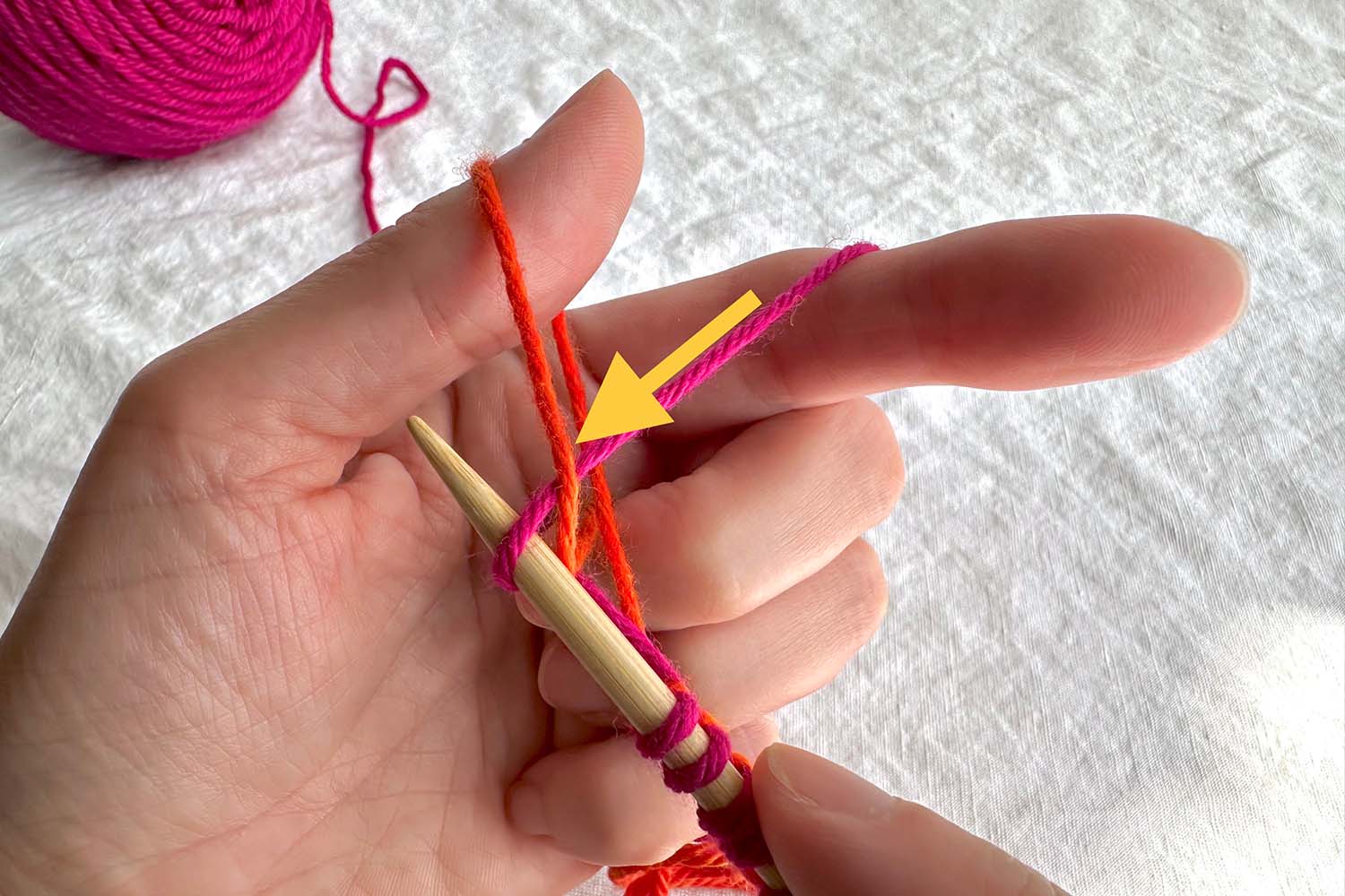 Needle pulling loop of yarn through thumb loop with directional arrow.