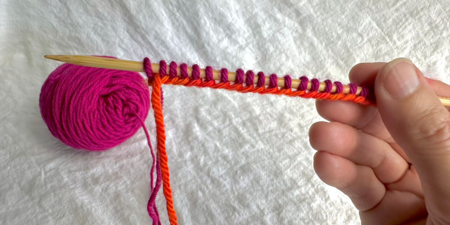Right side view of several pink stitches cast onto needle and base of stitches shows the path of the orange tail yarn.
