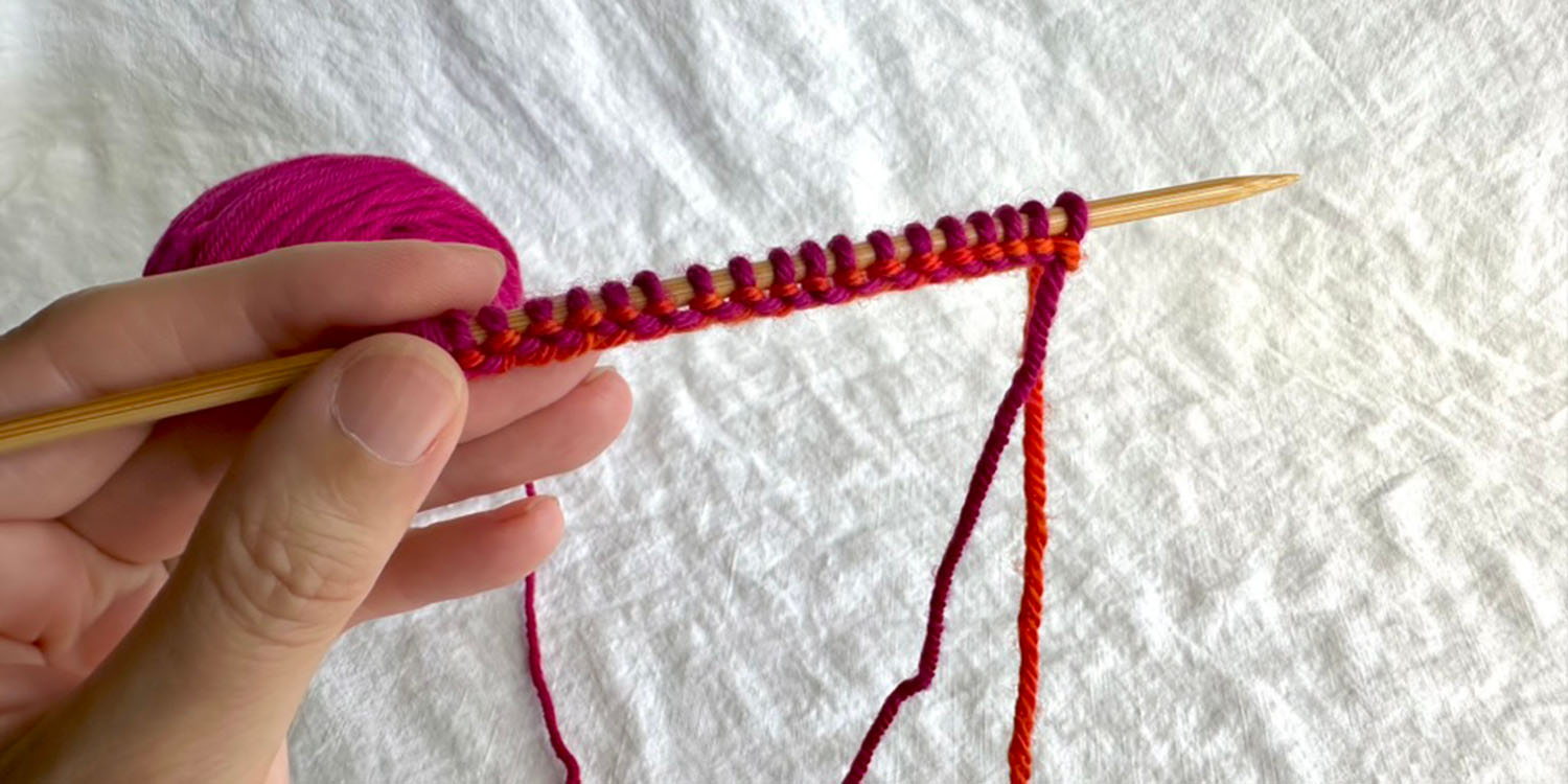 Wrong side view of several pink stitches cast onto needle and base of stitches shows the path of the orange tail yarn.