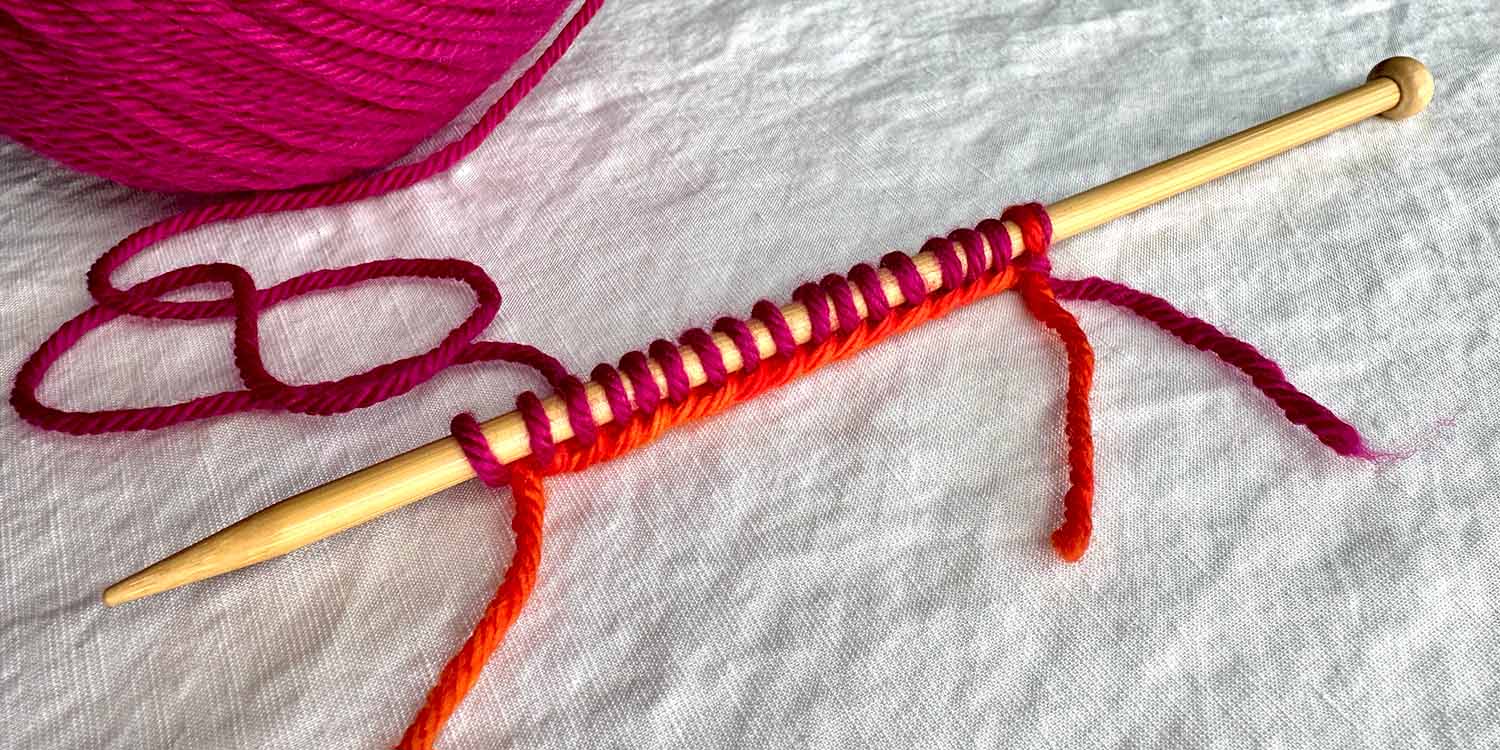 Several stitches cast onto needle started with a slip knot holding two strands together; orange used for tail and pink used for stitches.