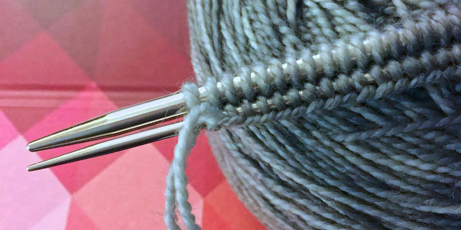 A cast on just completed on two needles (one larger, one smaller), sitting on top of a grey yarn cake, sitting on an iPad cover with red and pink triangles.