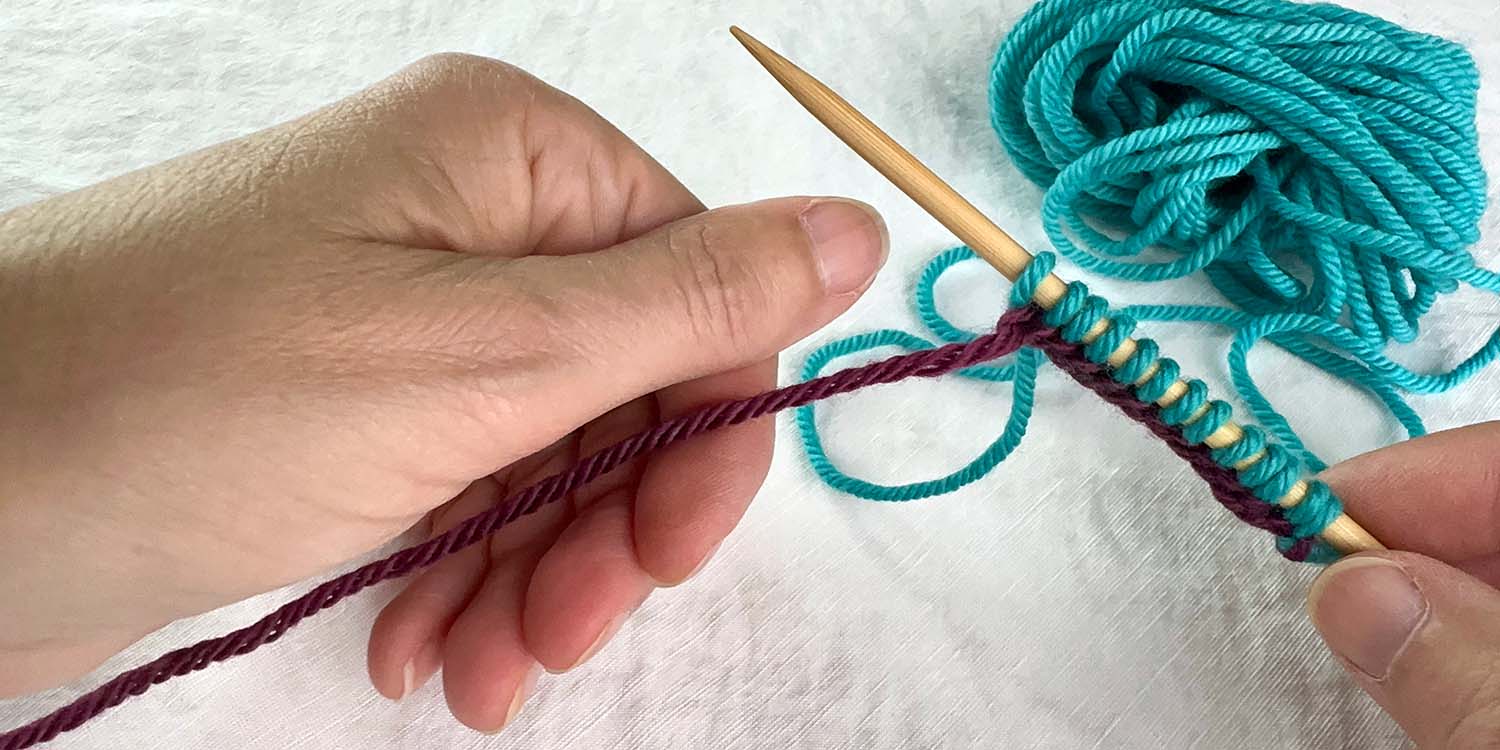 Newly cast on blue stitches on needle held in right hand and purple tail yarn being grasped by left hand, palm down with thumb above yarn.