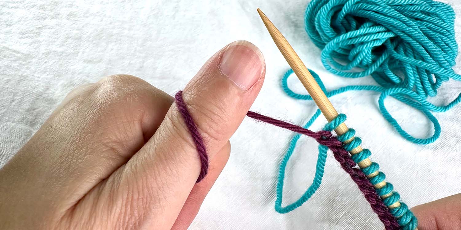 Newly cast on stitches on needle held in right hand while left hand grasps tail yarn which is wound around left thumb which is pointing upwards.