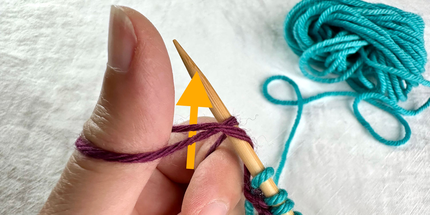 Left thumbs pointing up with a loop of yarn wrapped around it. Knitting needle inserted underneath both strands of loop and a directional arrow also illustrates path of needle.