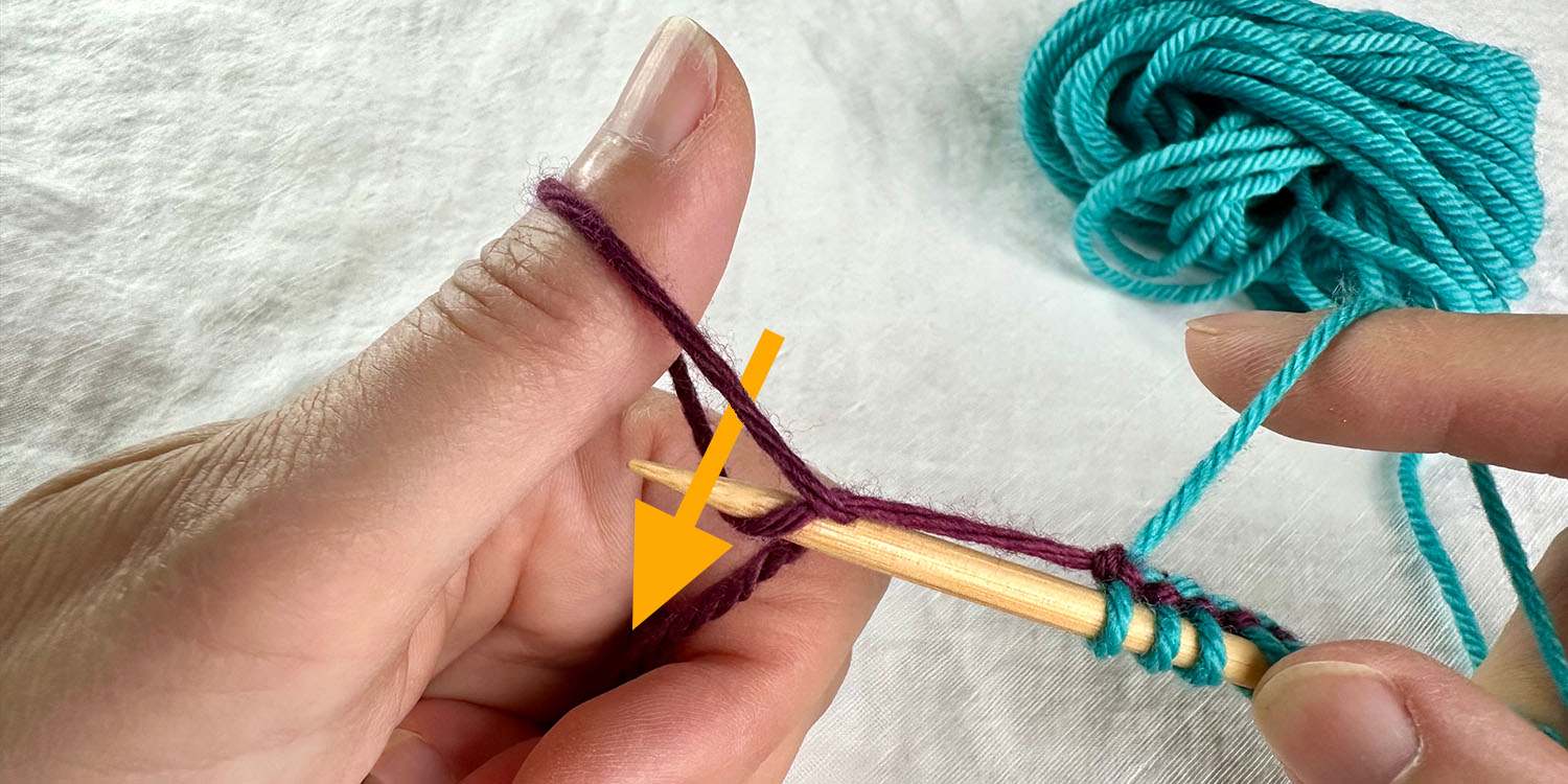 Needle held in right hand with cast on stitches has been inserted into loop on left thumb from above. Directional arrow illustrates path of needle.