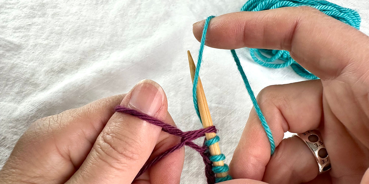 Needle in right hand with blue stitches plus purple loop from yarn wrapped around left thumb, then right forefinger is wrapping blue yarn aroud needle as if to knit.