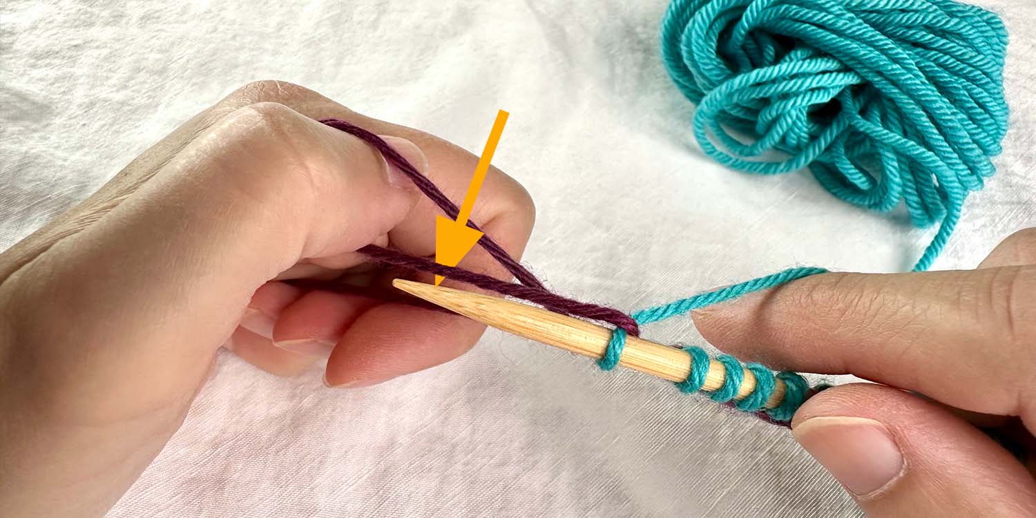 Needle in right hand and loop of yarn around left thumb. Needle has moved through centre of loop in a downwards motion as indicated by a directional arrow.